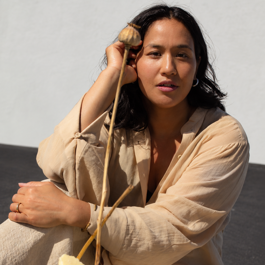 A woman in a beige linen outfit sits outdoors next to a large metal bowl containing pale yellow handmade soap bars, with dried flower buds hanging near her head.
